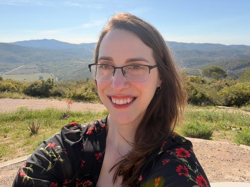 Klaudia Warner, smiling, wearing glasses and a black dress with red flowers, on a sunny hilltop with green hills behind her