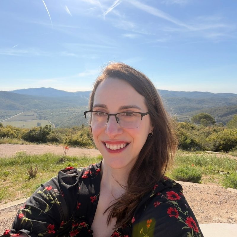 Klaudia Warner, smiling, wearing glasses and a black dress with red flowers, on a sunny hilltop with green hills behind her