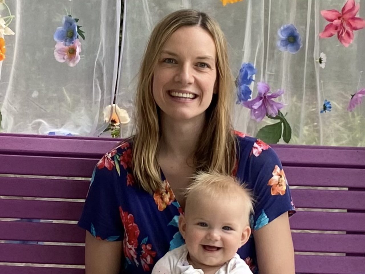 Patrycia Leja, smiling, on a purple bench with her baby daughter Lena on her lap, in front of a sheer curtain decorated with colorful flowers